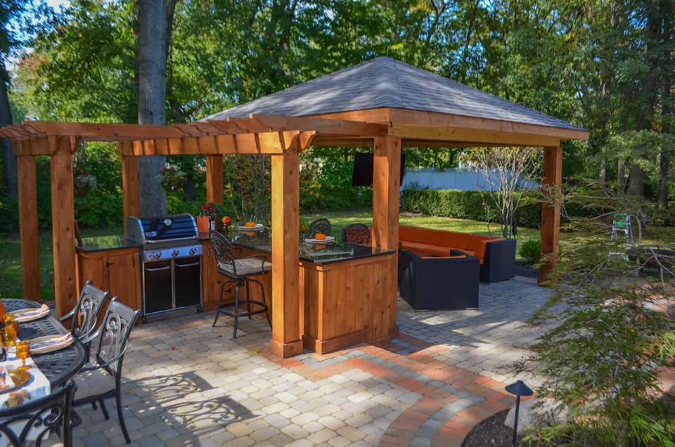 Backyard pavilion and outdoor kitchen with paver patio, dining table, lounge seating, and fall trees.